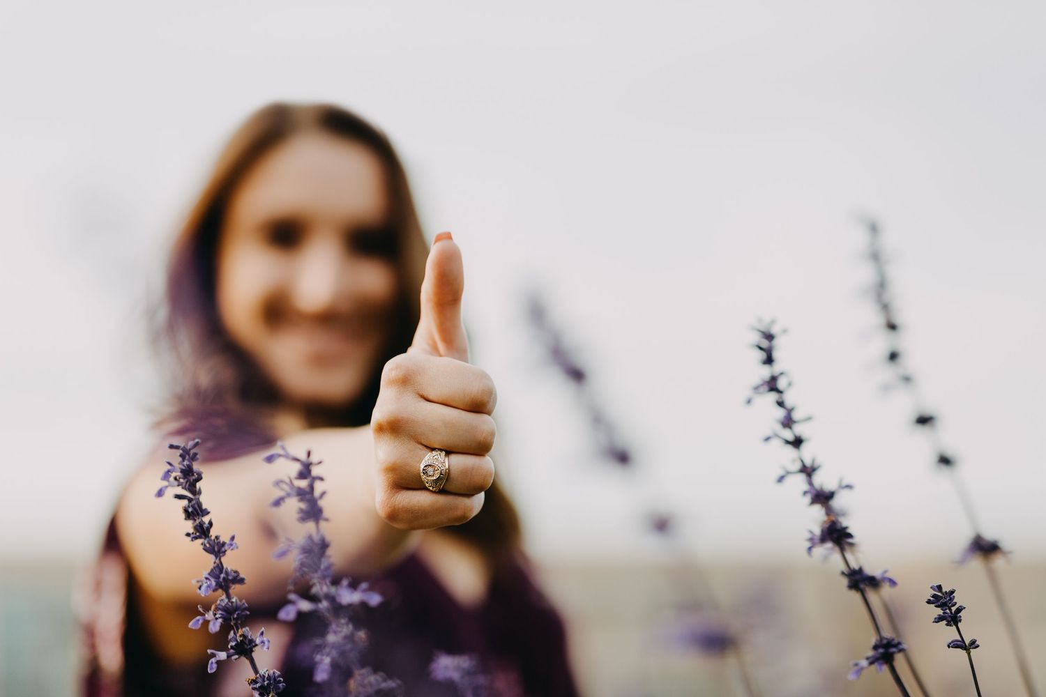 Woman in purple dress giving thumbs up among lavender flowers, wearing a diamond ring, smiling outdoors.