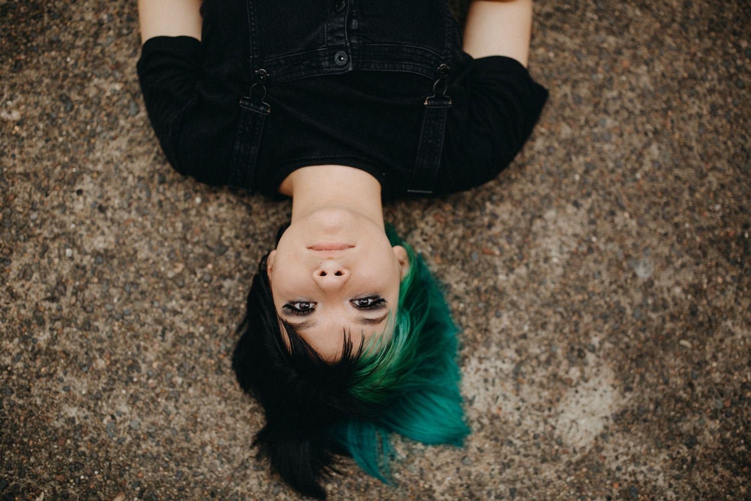 Young woman with teal and black hair lying on ground, wearing black outfit, viewed from above.