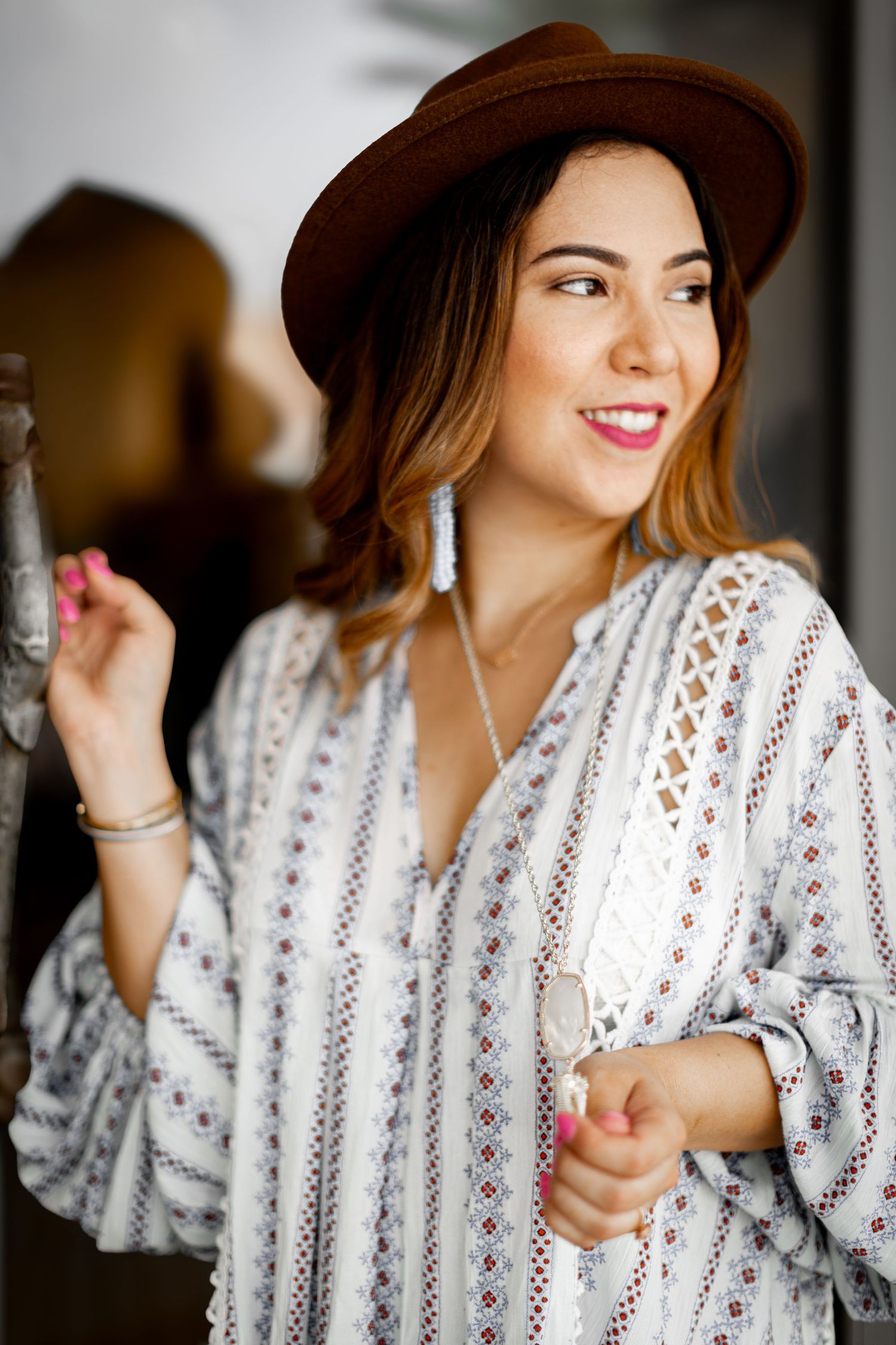 A smiling woman in a boho patterned blouse and brown hat poses with dangling earrings.
