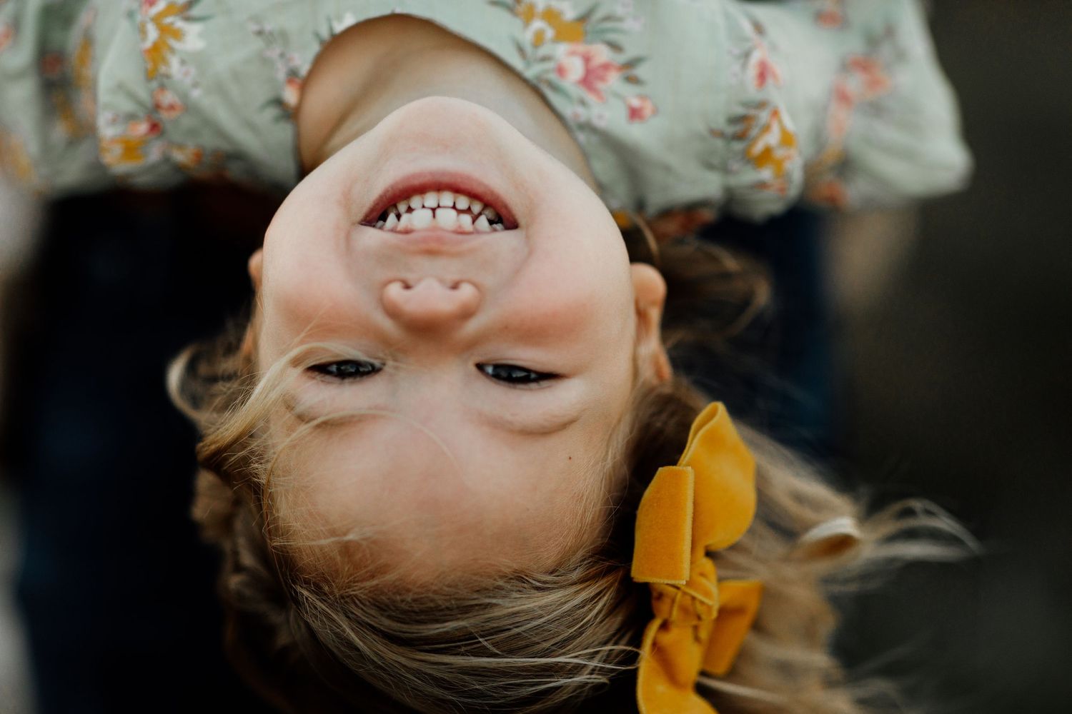 A joyful young girl with blonde hair laughs upside down, wearing a floral top and yellow bow.
