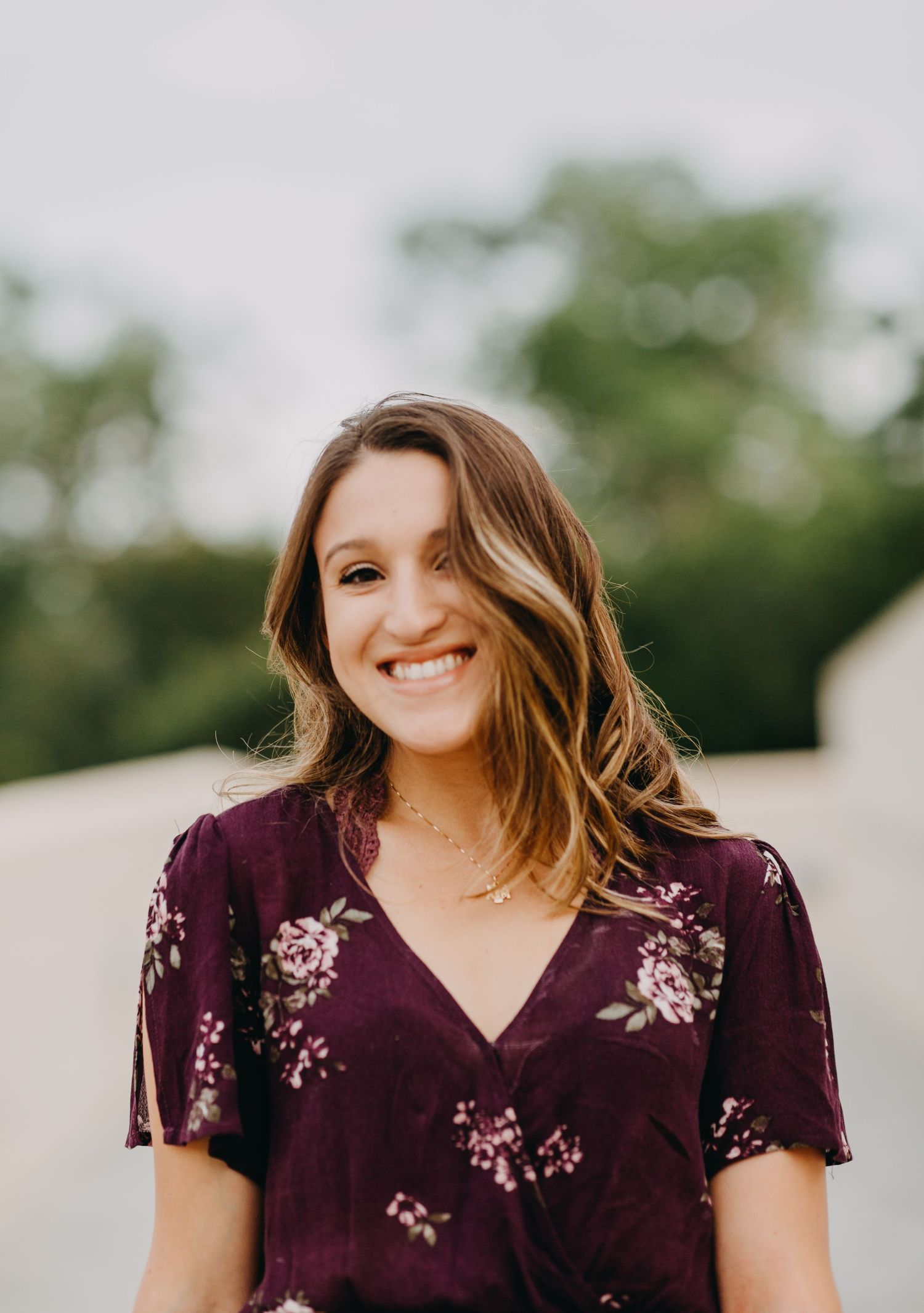 Young woman smiling outdoors in a purple floral dress with lush green trees in the background.