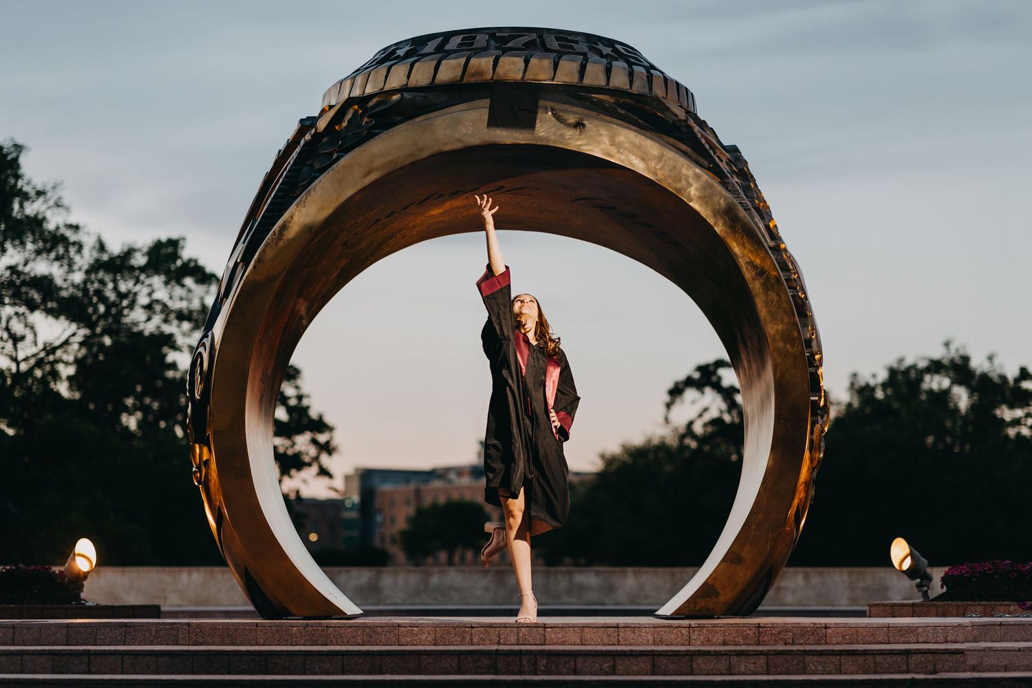 A dancer poses gracefully inside a large golden circular sculpture at dusk with trees in the background.