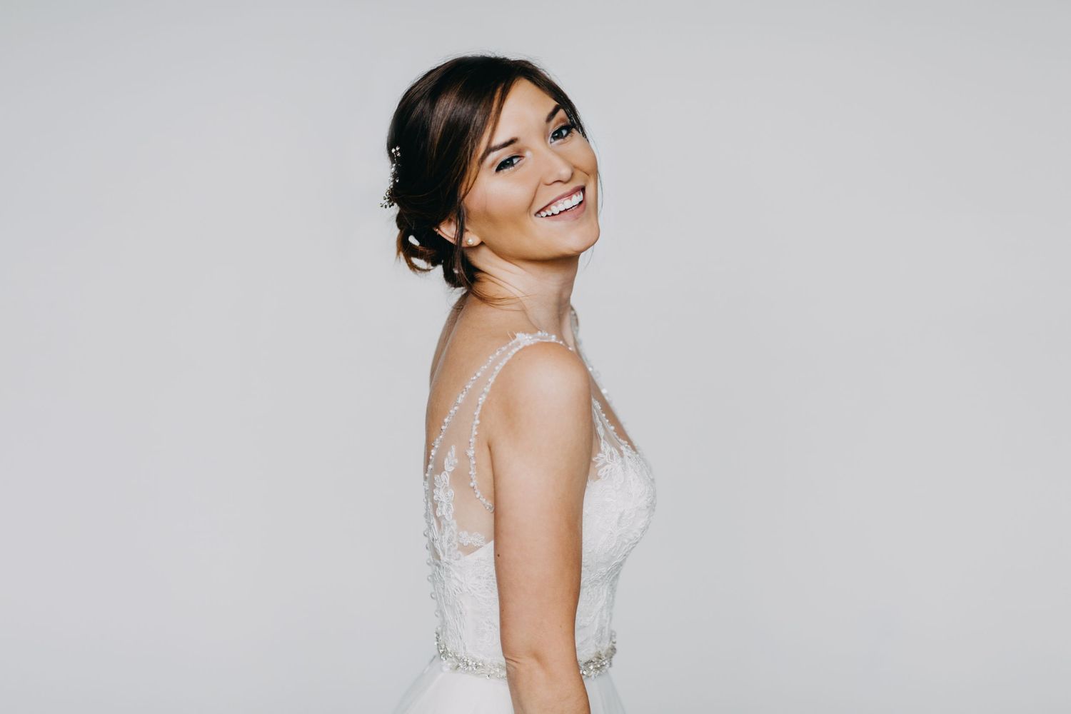 Smiling bride in elegant white lace gown posing against a light gray background.
