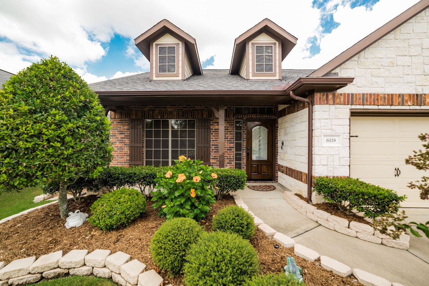 Charming brick home exterior with dormer windows, manicured shrubs, and blooming flowers.