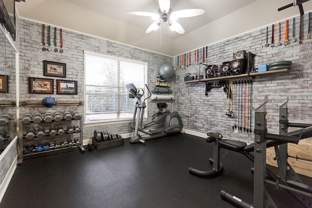 Home gym with brick walls, weight racks, elliptical machine, and rubber flooring with ceiling fan.