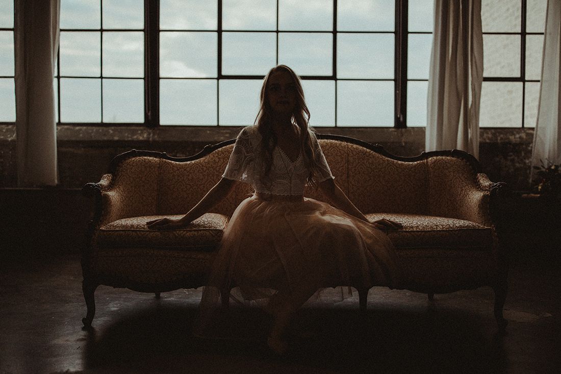 Bride in white gown sitting on vintage gold sofa before large industrial windows in moody light.