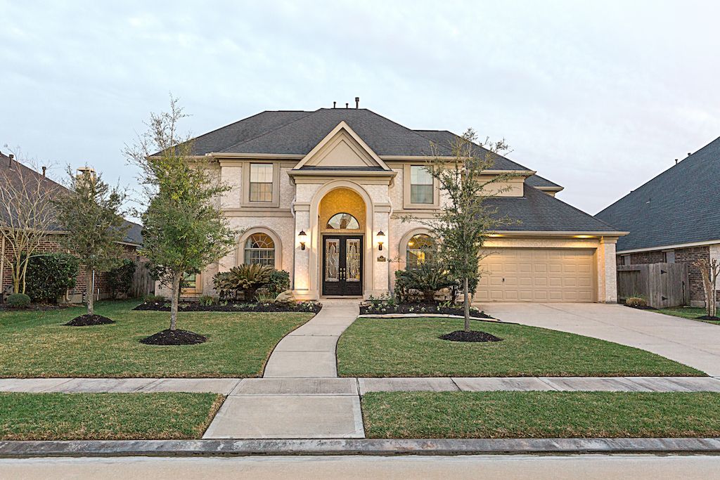 Elegant two-story brick home with arched entryway, three-car garage, manicured lawn, and mature trees in a suburban neighborhood.