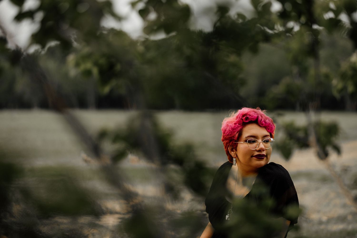 Woman with vibrant pink hair wearing black outfit laughing outdoors among green trees and fields.