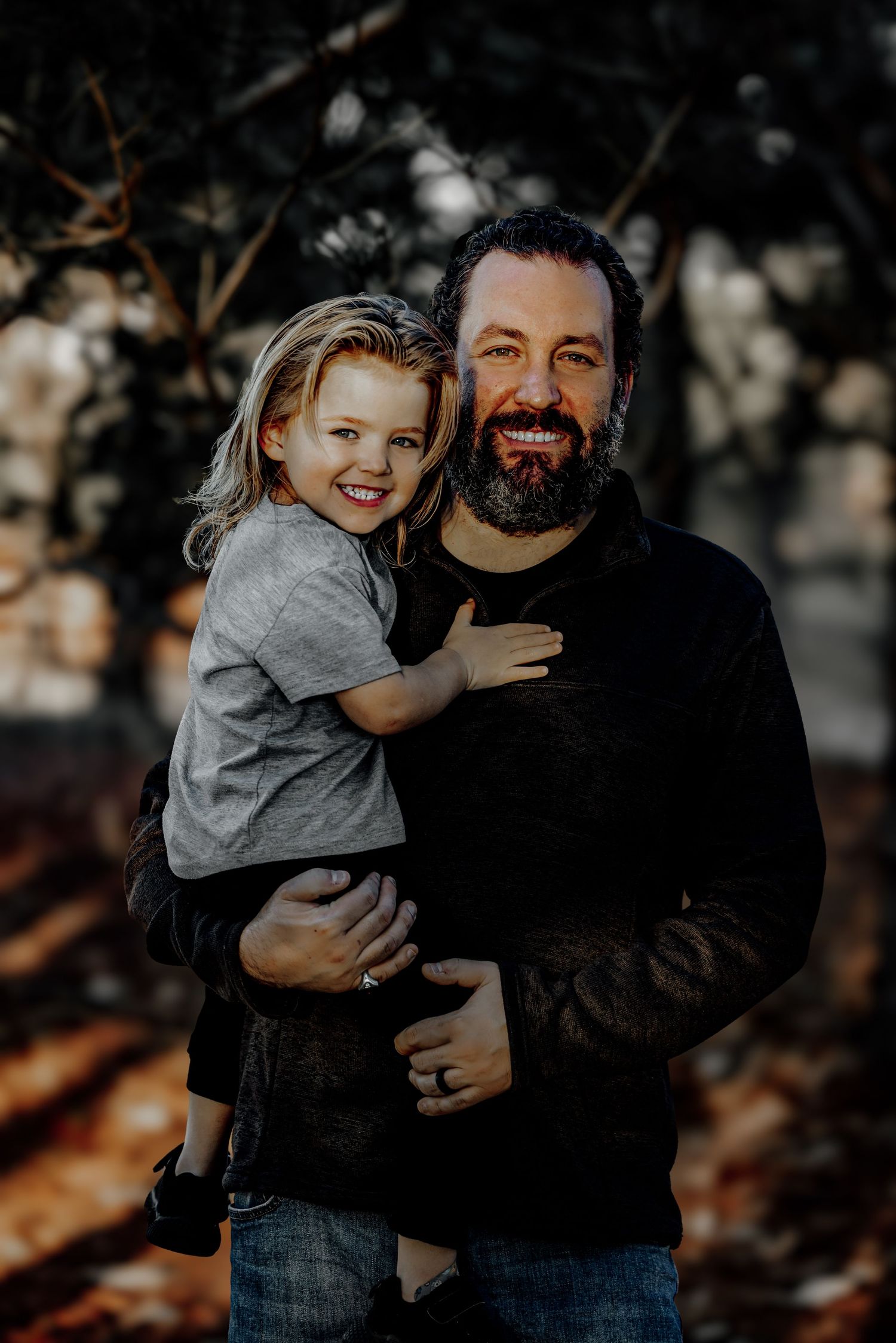 A bearded man in a dark sweater holds a smiling blonde child in a sunlit outdoor fall setting.