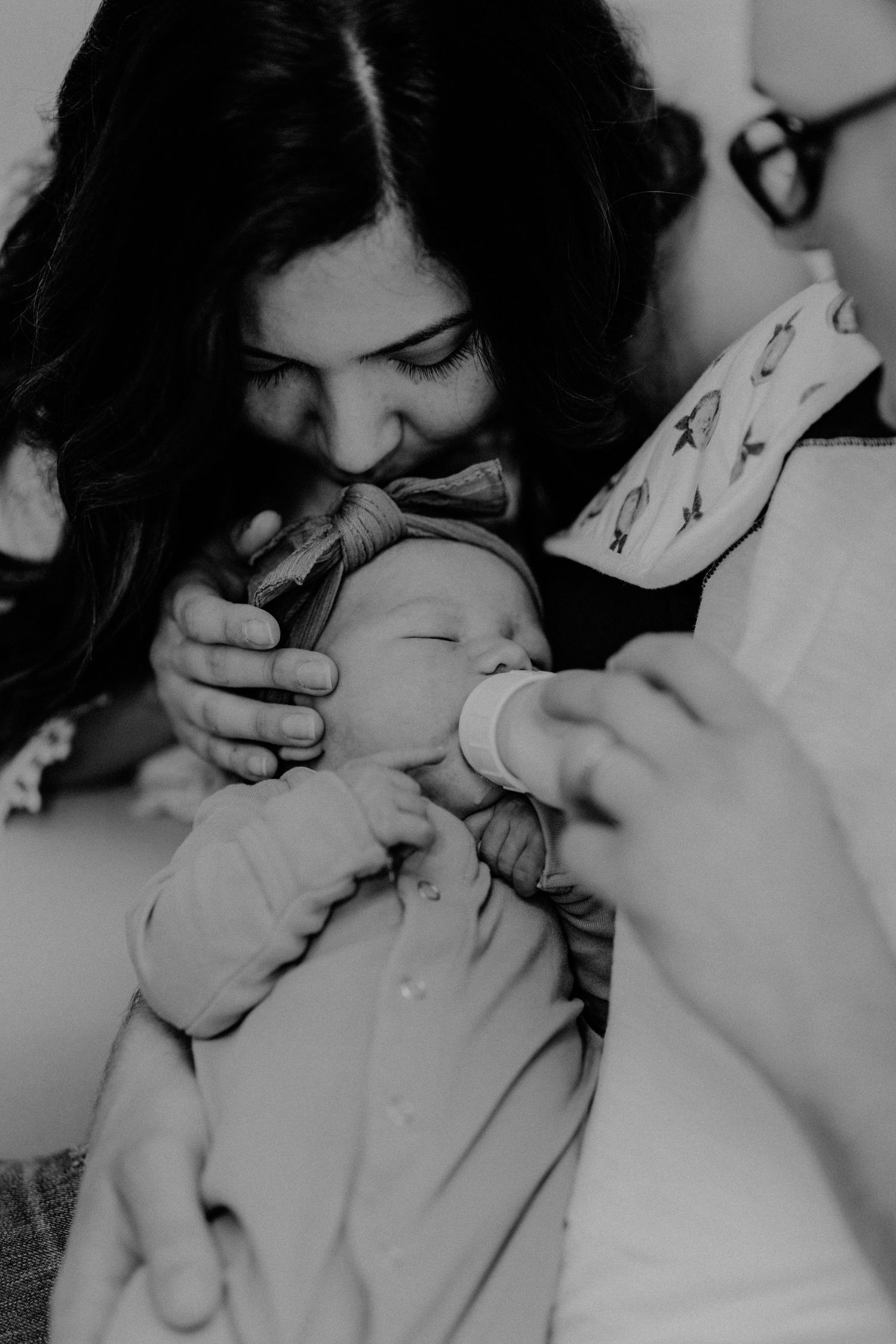 Black and white photo of mother tenderly kissing newborn baby while bottle feeding.