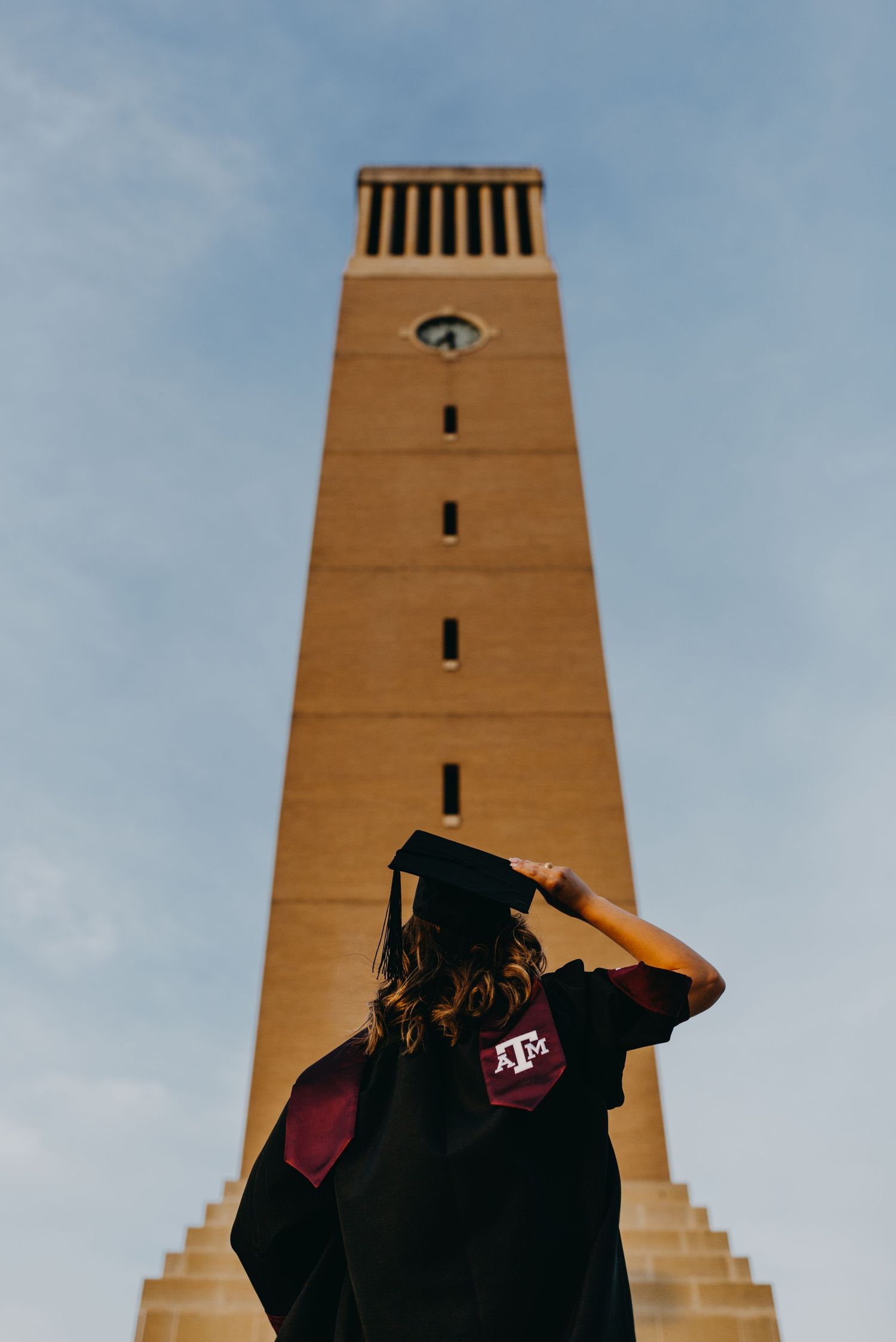 Texas A&M graduate in cap and gown stands before the iconic Albritton Bell Tower on a partly cloudy day.