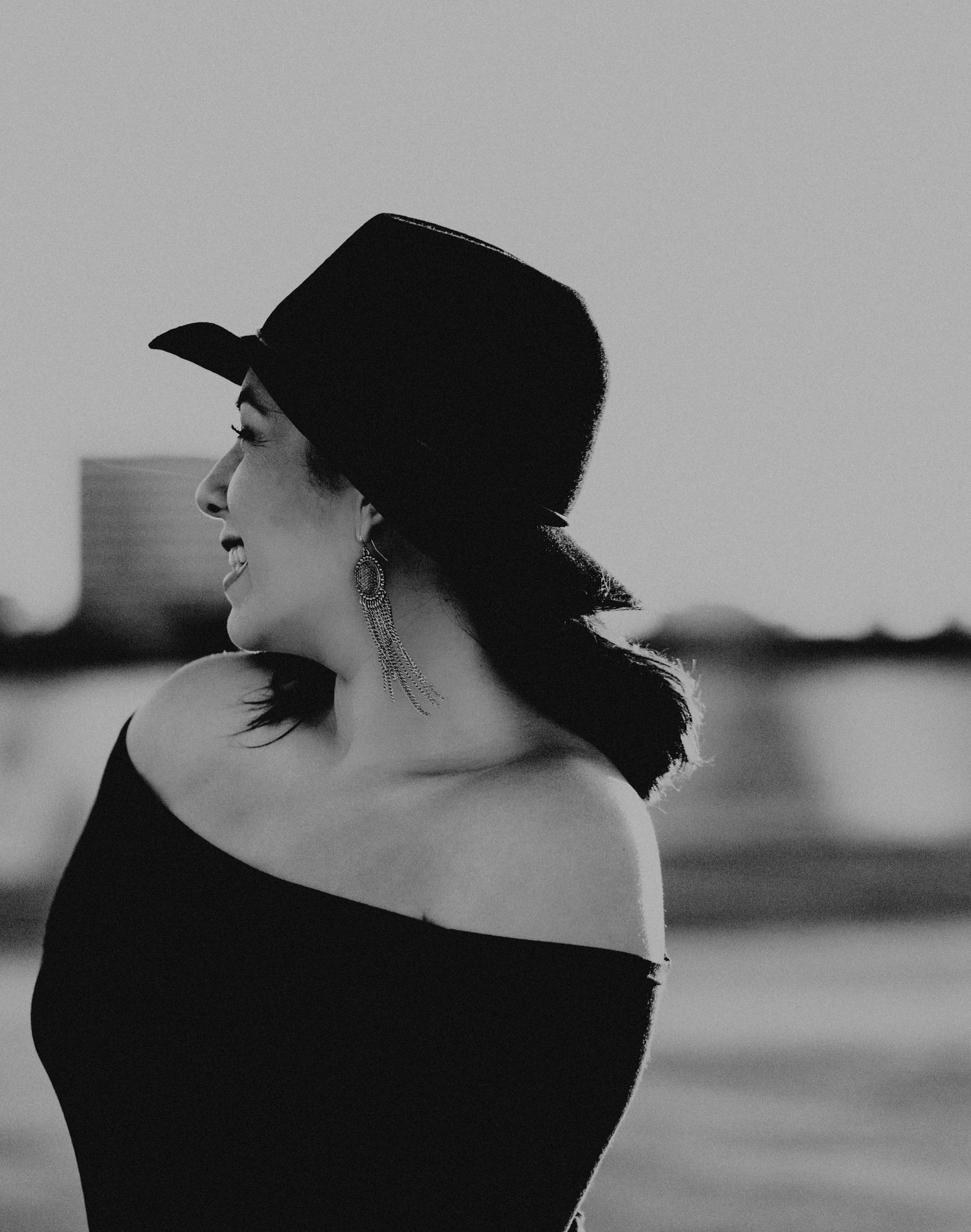 Black and white photo of a woman in an off-shoulder top and wide-brim hat, gazing sideways near a waterfront.
