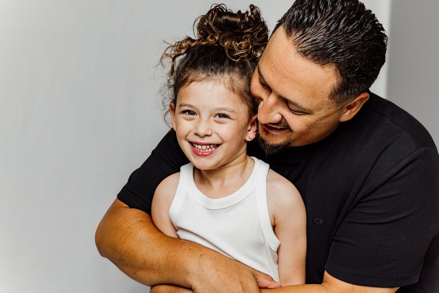 A father hugging his smiling young daughter in a warm, candid portrait against a light gray background.