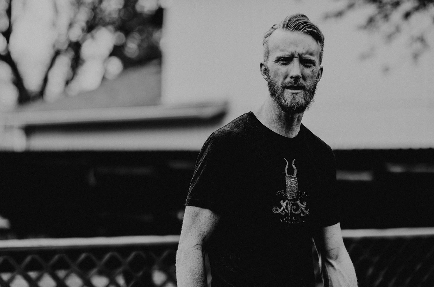 Black and white portrait of a bearded man in a black graphic t-shirt standing outdoors.