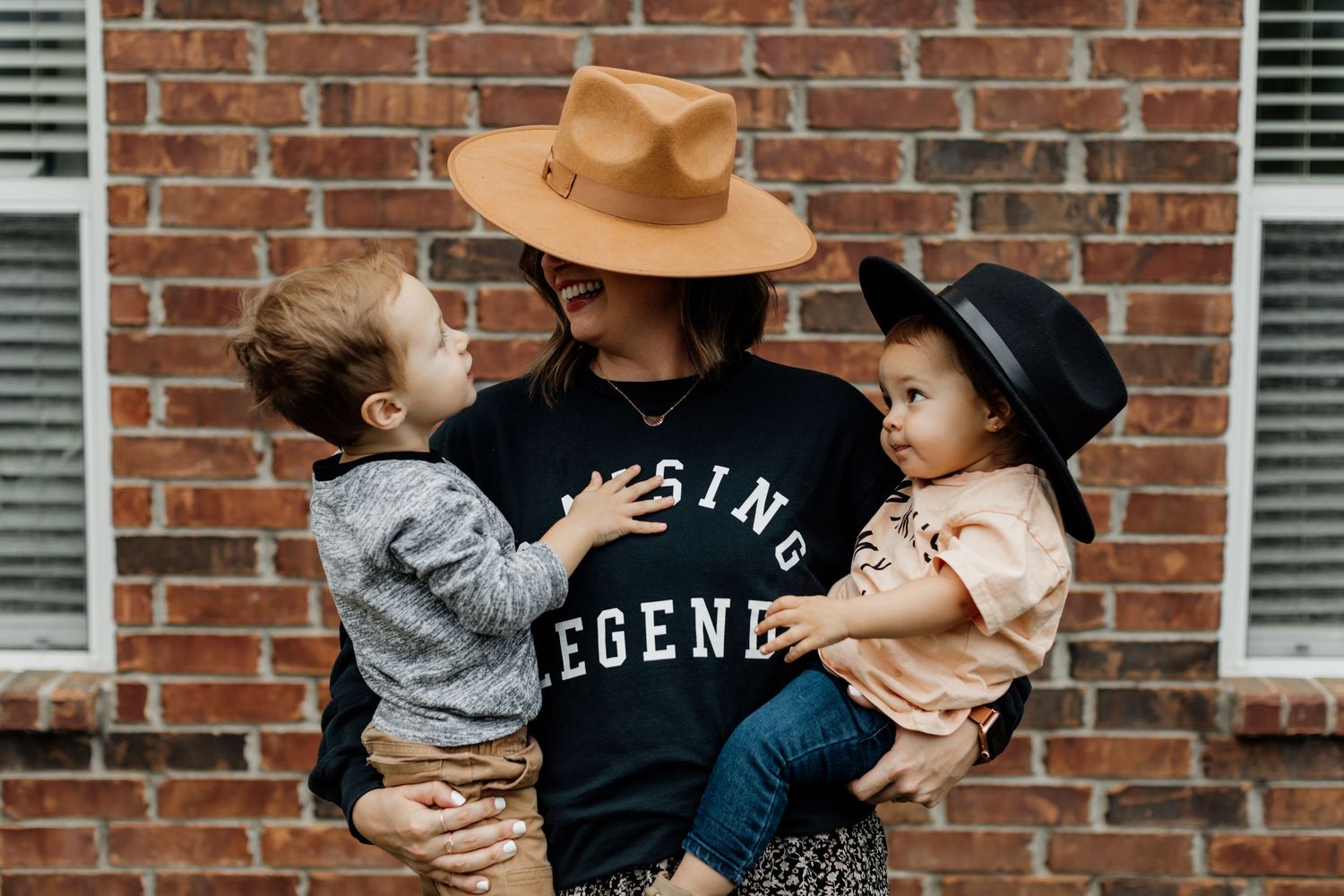 Person in black graphic tee and tan hat holding two toddlers wearing hats in front of a brick wall.