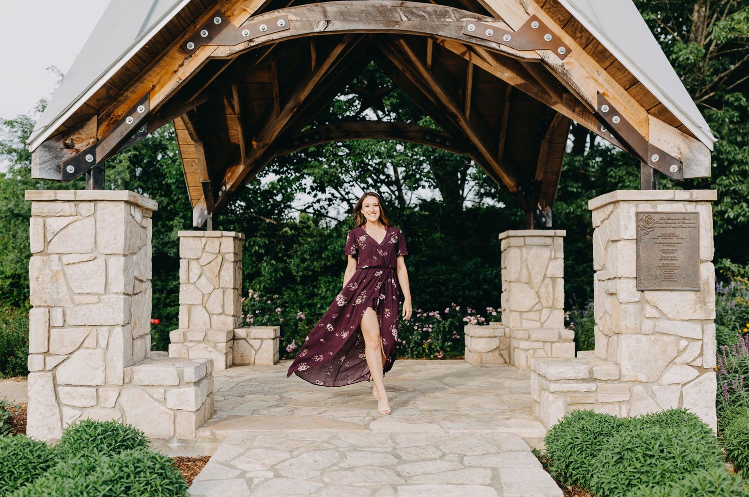 Woman in floral burgundy wrap dress walking under stone pavilion with wooden roof surrounded by lush greenery.