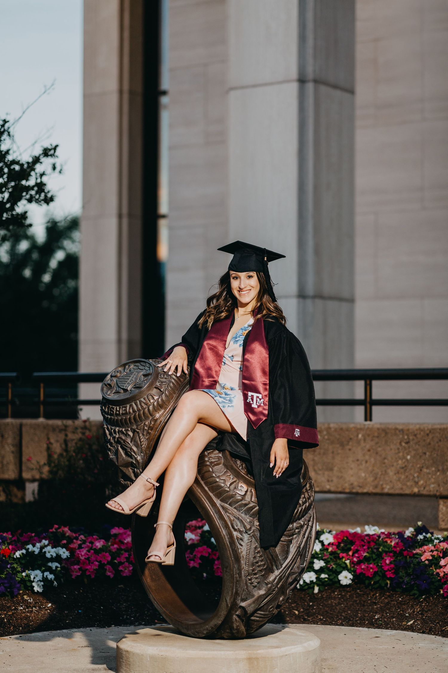 Smiling graduate in cap and gown poses on a bronze mascot statue with colorful flowers and columns behind her.
