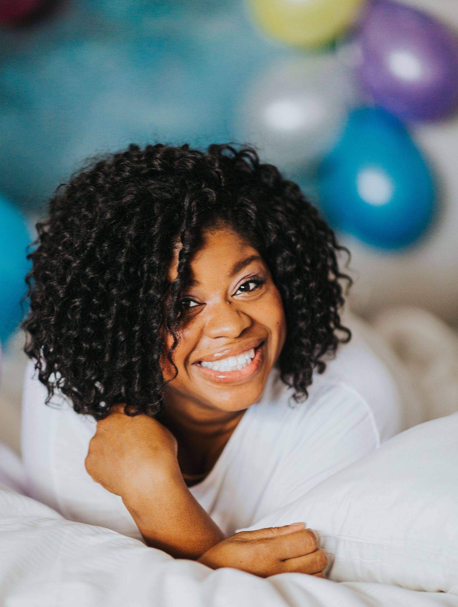 A smiling young Black woman with curly hair lying on a bed surrounded by colorful balloons.