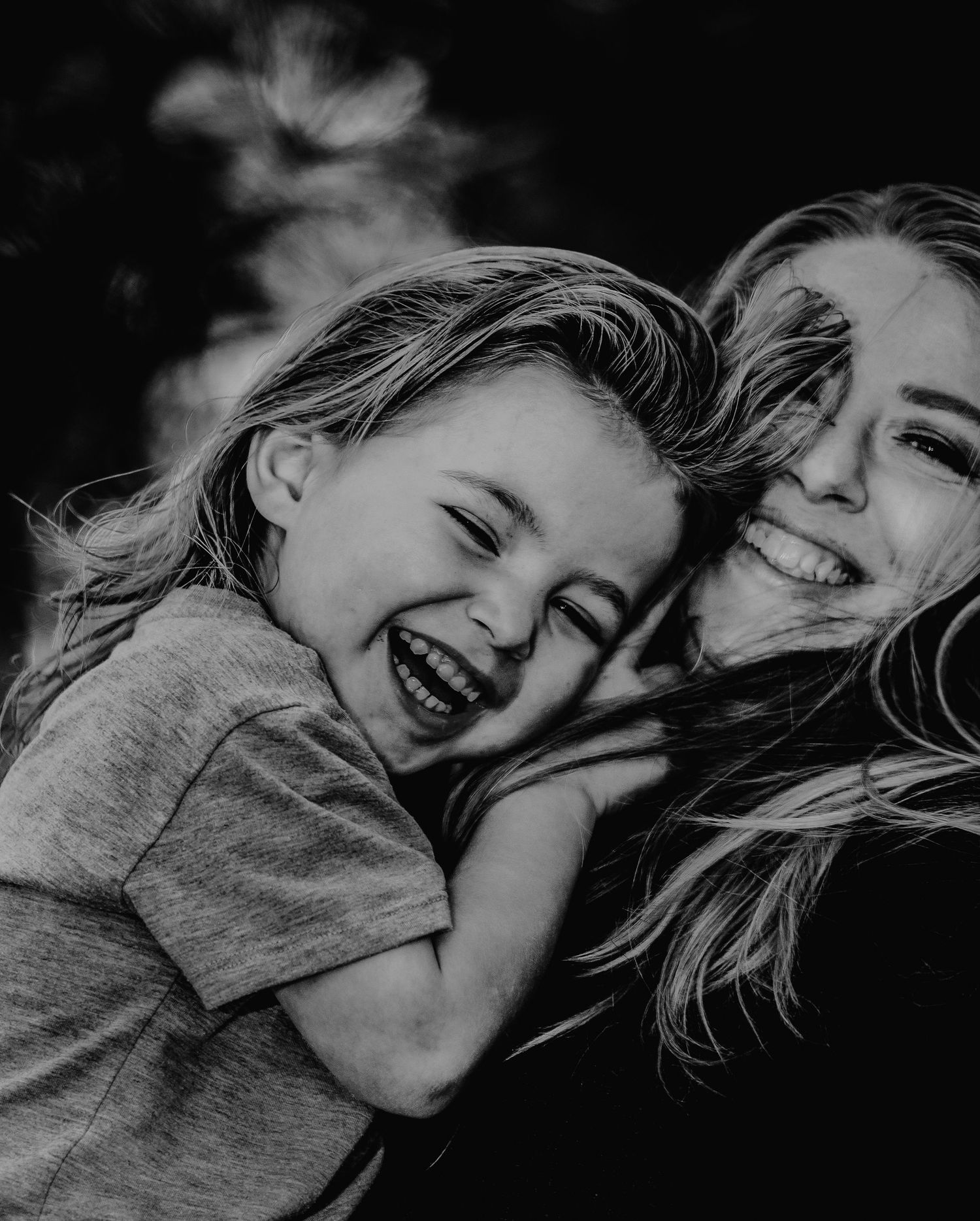 Black and white photo of a laughing child embracing a smiling woman in a joyful outdoor portrait.