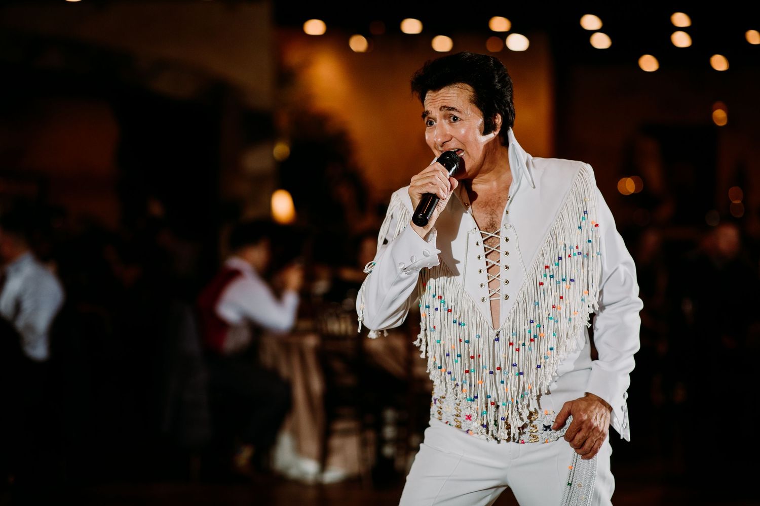 Elvis impersonator performing in white jeweled jumpsuit at an indoor wedding reception.
