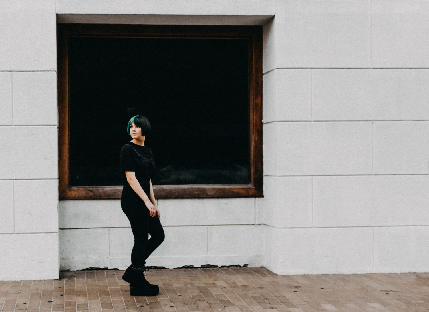 Young woman with teal hair in all-black outfit posing against white brick wall with dark window.