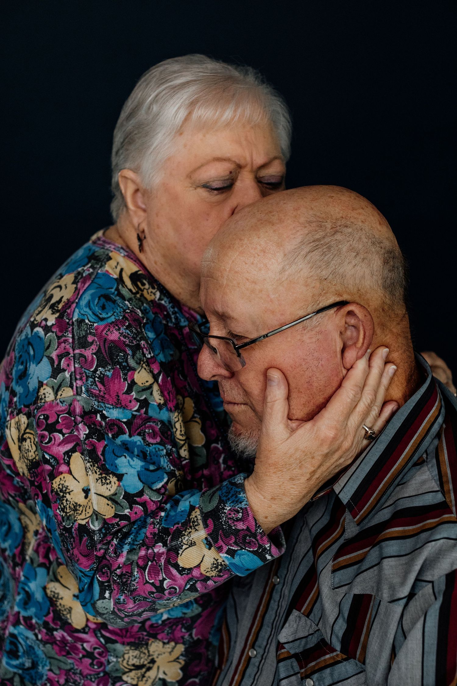 Elderly woman tenderly embracing and kissing her husband against dark background.