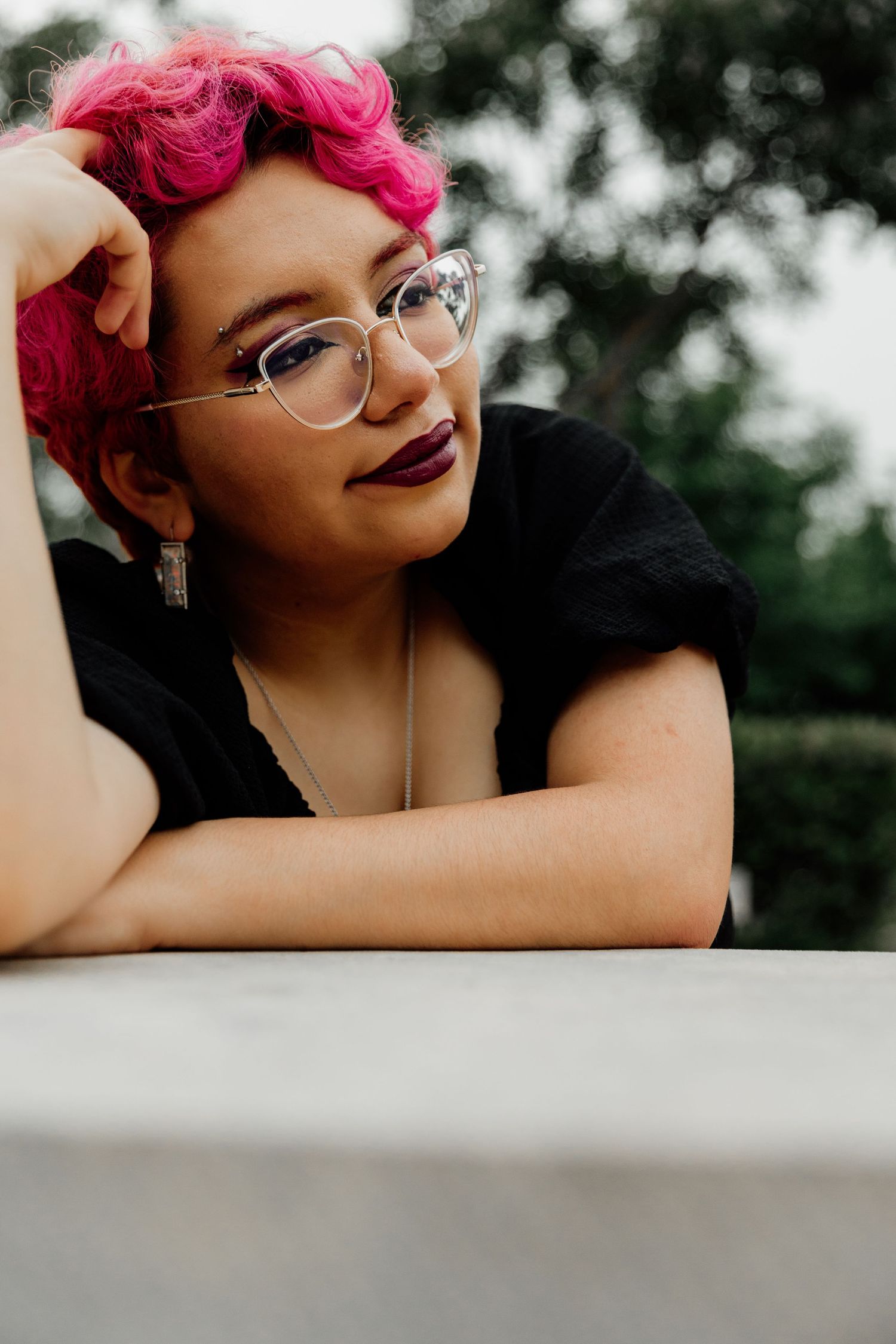 Young woman with pink hair and clear glasses wearing a black top, leaning on a ledge outdoors.