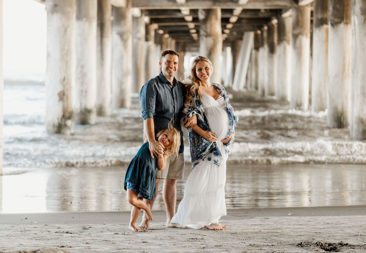 Happy family posing under a beach pier, with a pregnant woman in white dress and playful child.