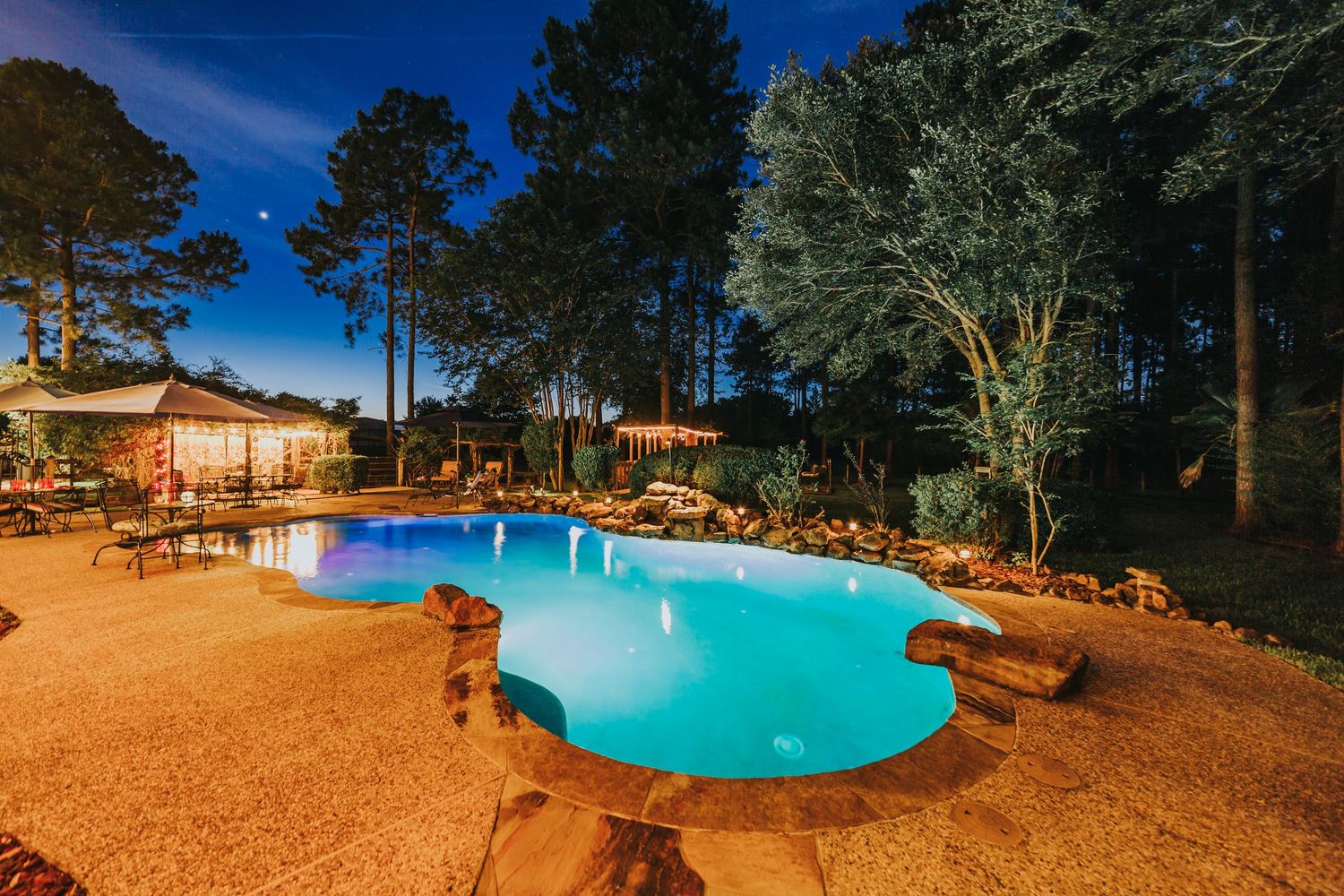 Stunning backyard pool illuminated at night surrounded by mature trees and patio.