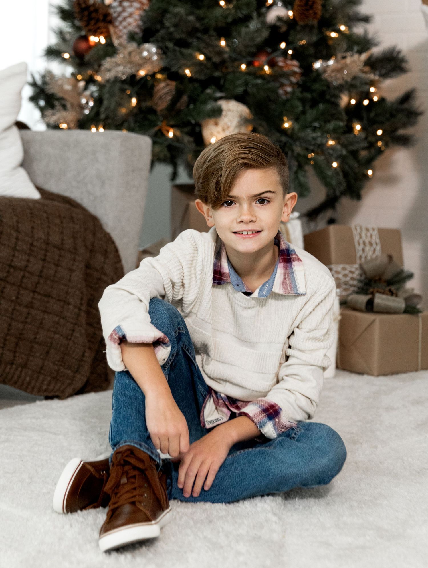 Smiling boy in cream sweater sitting cross-legged before a decorated Christmas tree.