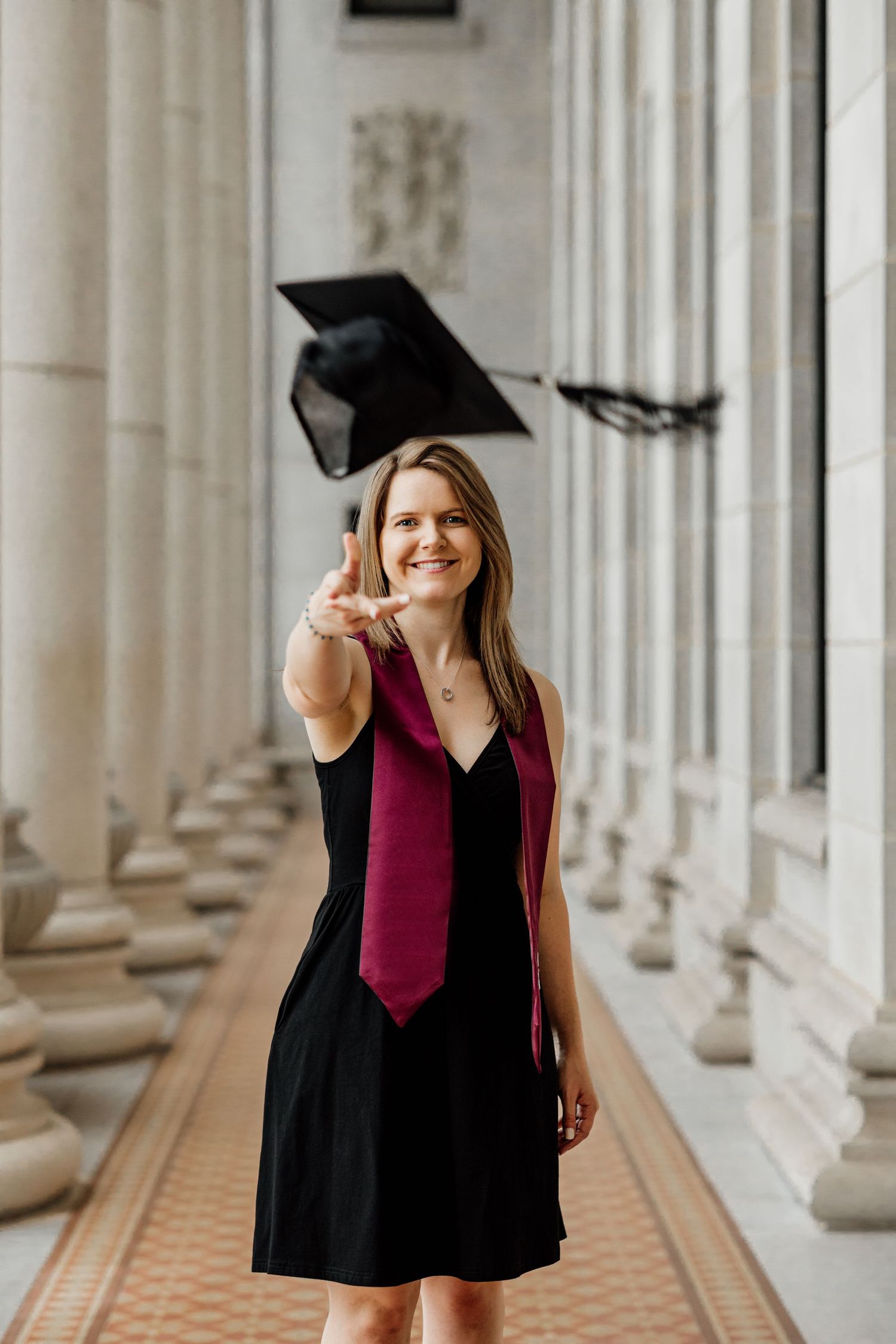 Graduate in black dress tossing cap while smiling between columns.