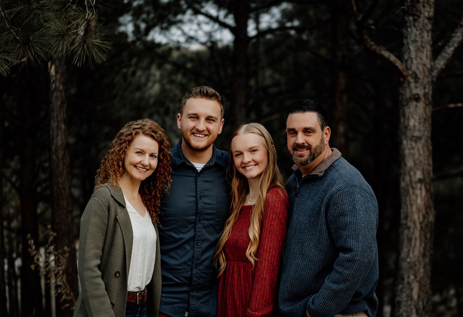 Family of four smiling together outdoors among pine trees.