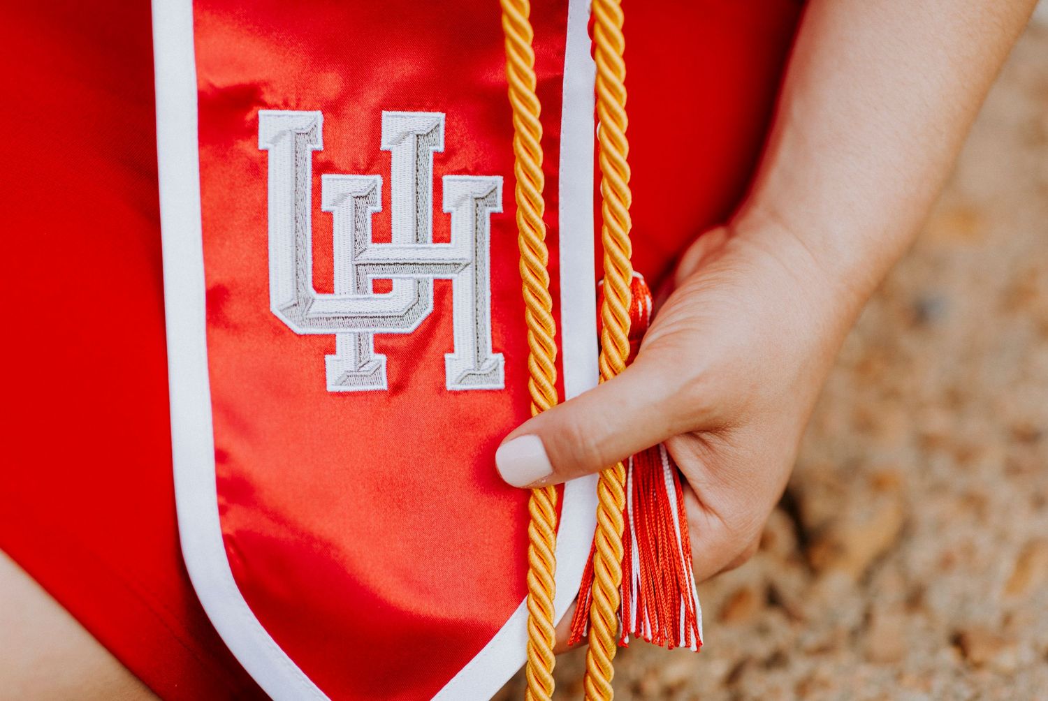 Close-up of University of Houston red graduation stole with UH logo and gold honor cord with red tassel.