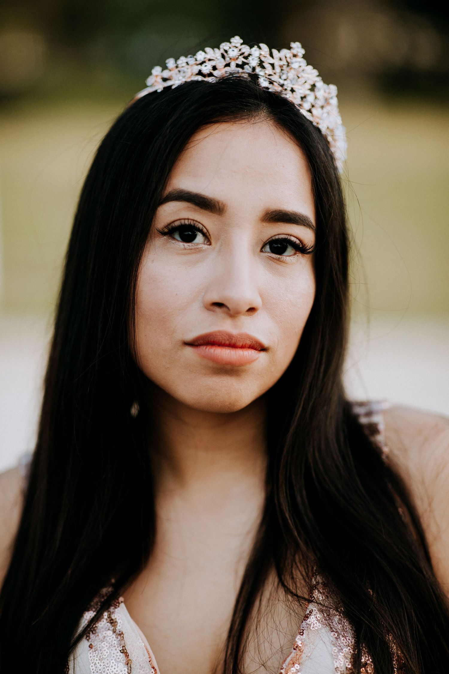 Young woman wearing a sparkling crystal tiara and sequined dress, with long dark hair and elegant makeup.