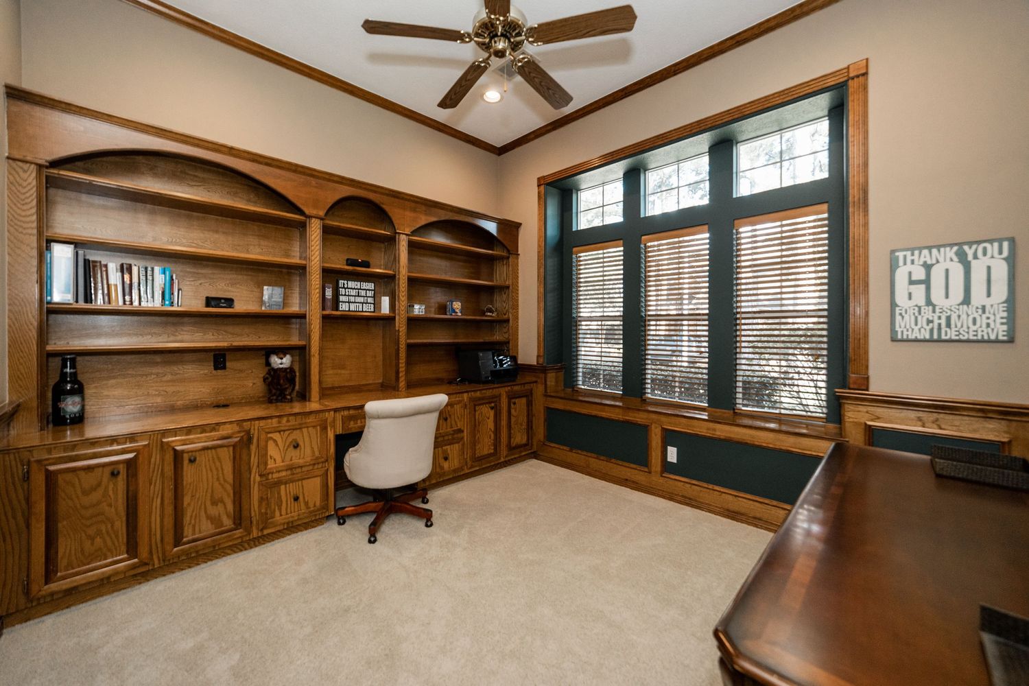Elegant home office with built-in oak shelving, a classic desk chair, large windows with blinds, and neutral carpet flooring.
