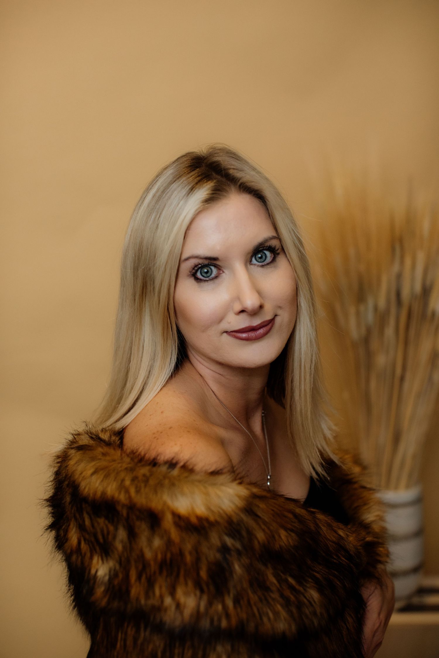 A blonde woman with blue eyes wearing a fur stole poses against a warm beige background.