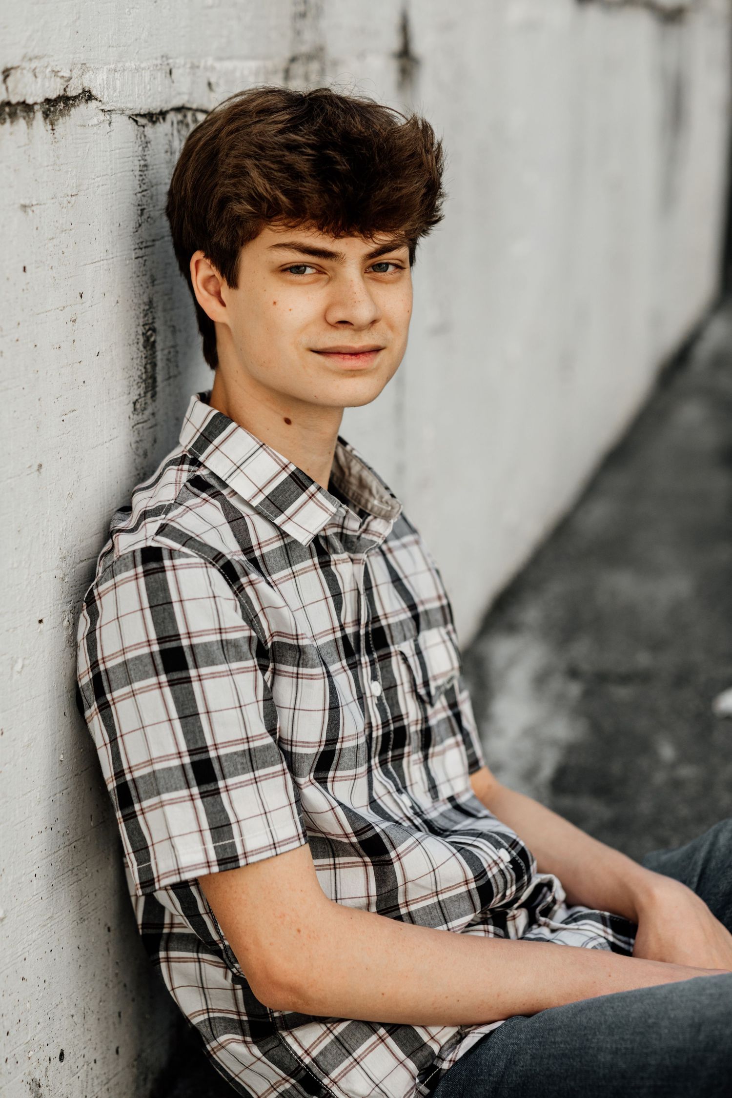 Young man in plaid shirt leaning against concrete wall, casual senior portrait photography.