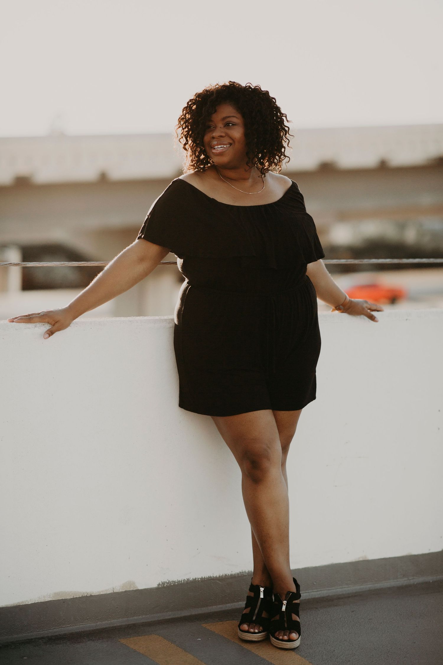 Confident Black woman in black off-shoulder dress posing on rooftop parking structure.