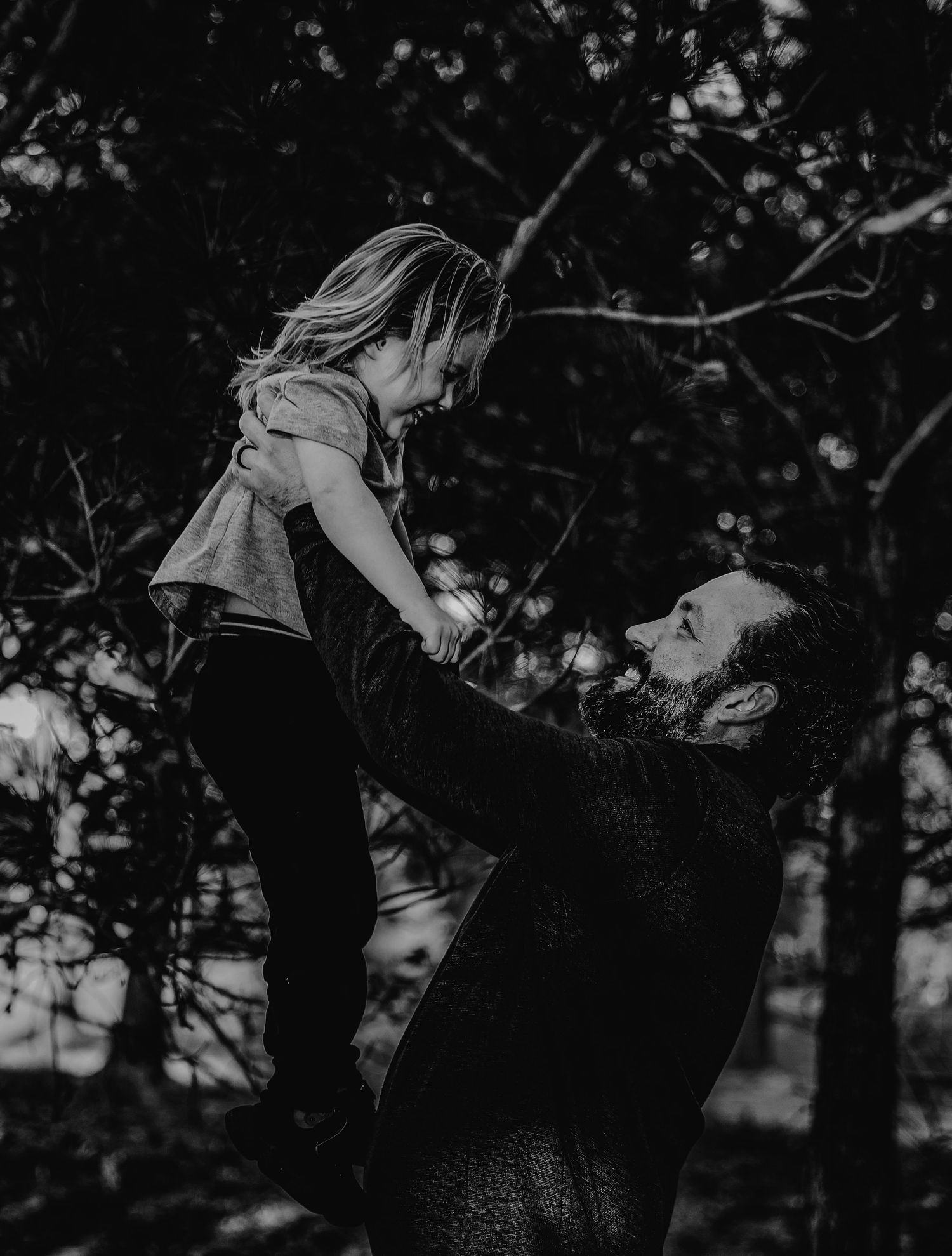 Black and white photo of a father lifting his young child high in the air among trees outdoors.