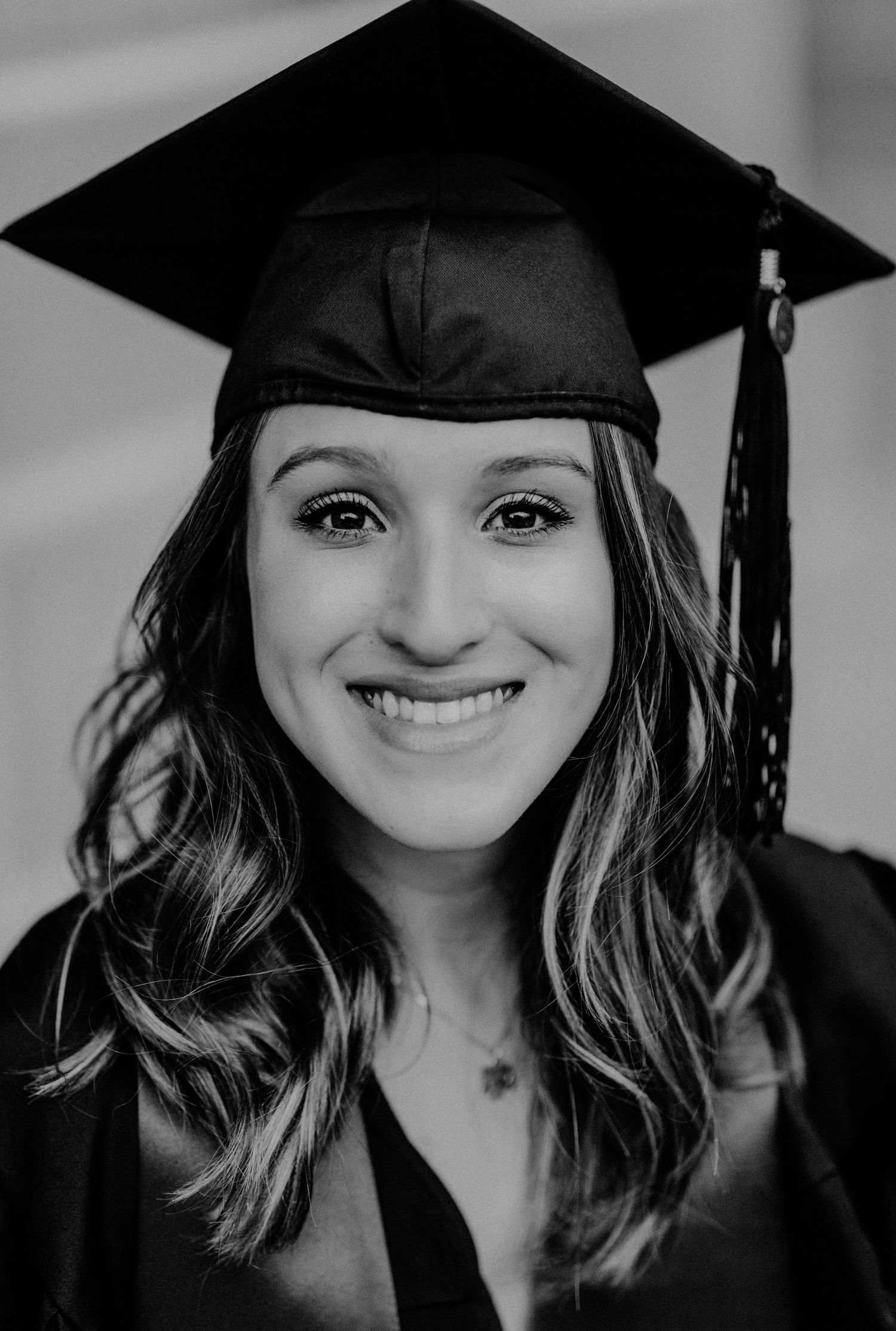 Smiling young woman in black graduation cap and gown posing for a portrait photo in black and white.