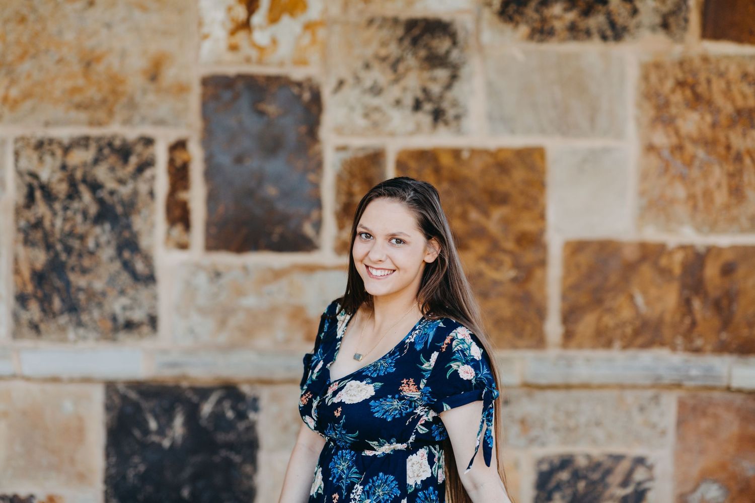 Smiling woman in floral dress posing against rustic stone wall outdoors.