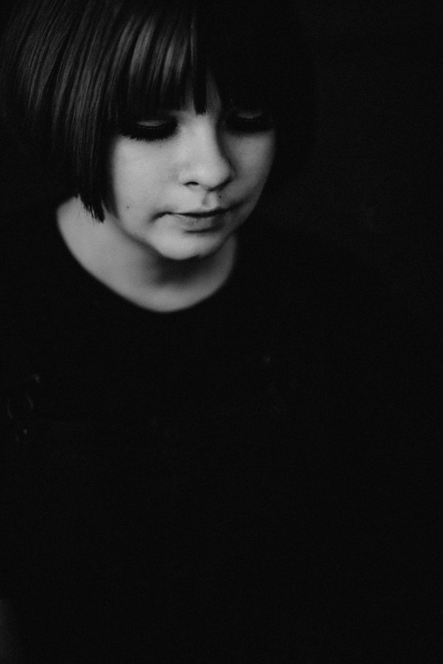 Black and white portrait of a child with a bob haircut looking down against a dark background.