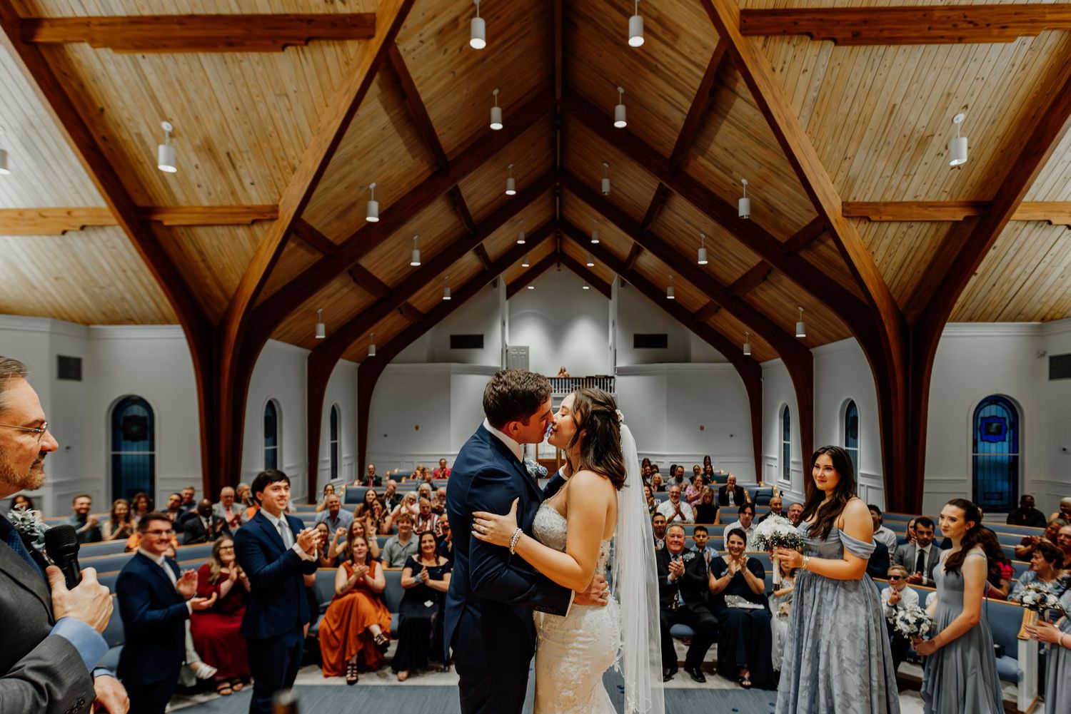 Bride and groom share first kiss at church altar as wedding guests cheer and applaud.