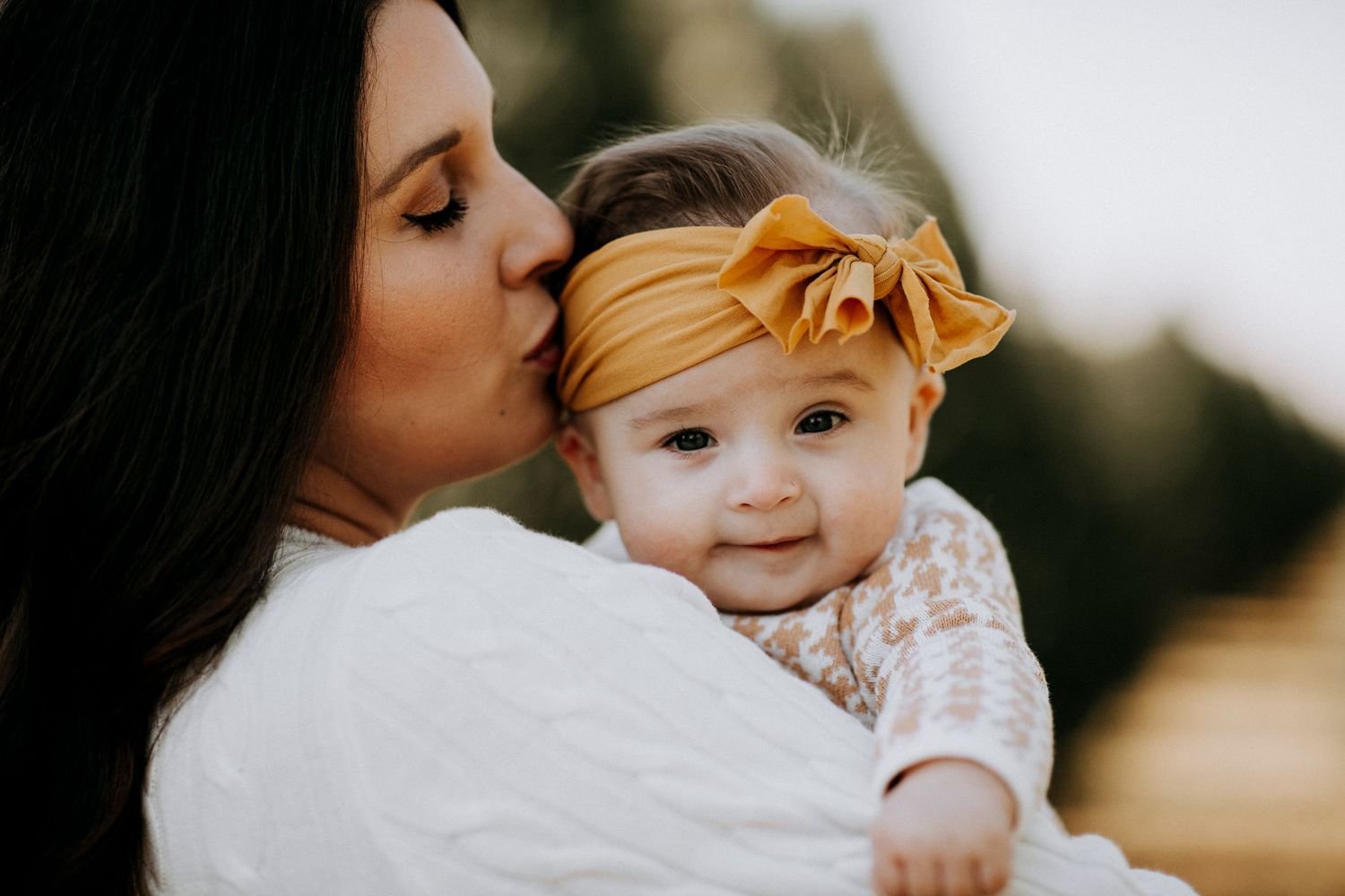 Mother gently kissing her baby girl with a mustard yellow bow headband.