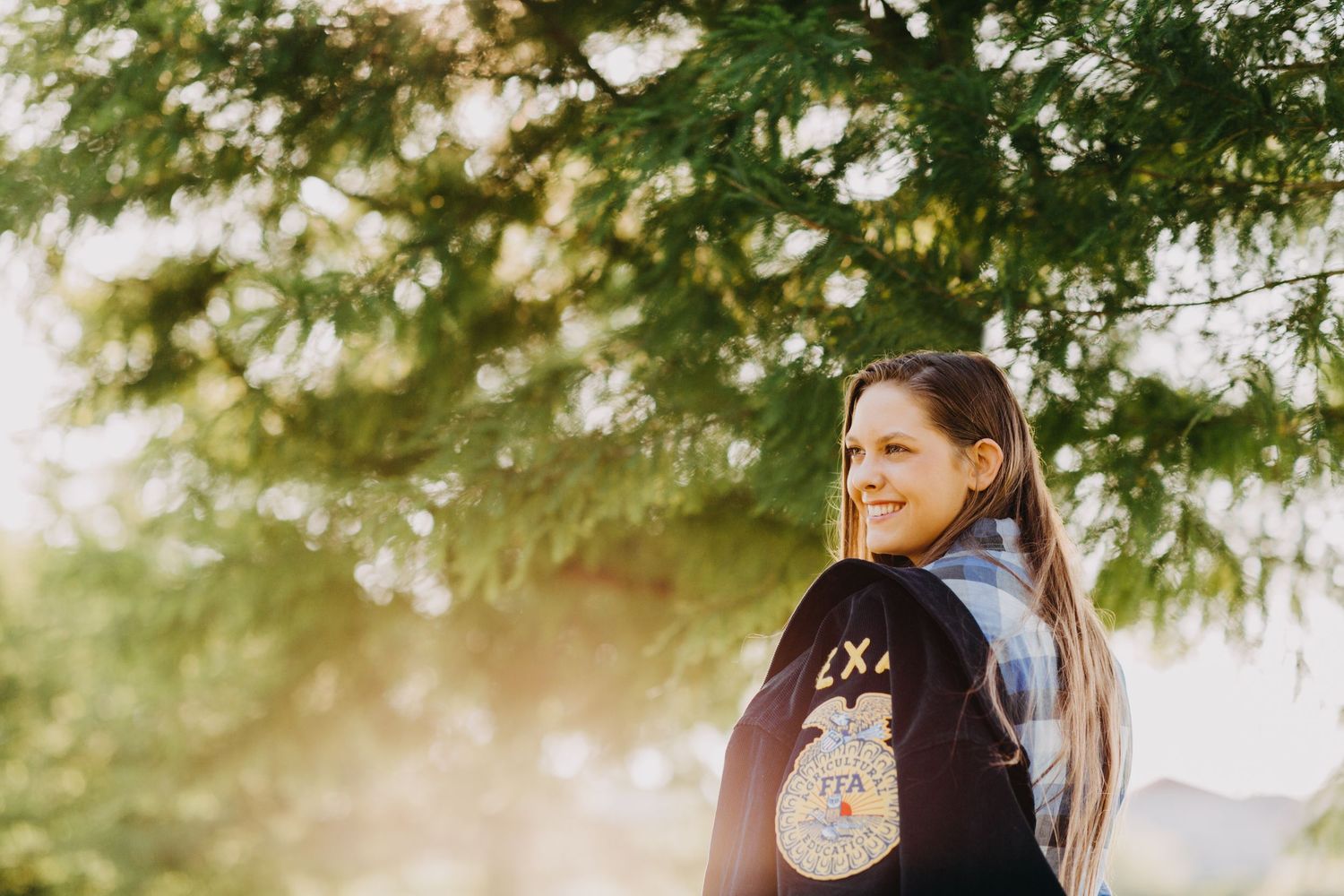 Young woman in FFA jacket smiling outdoors under green tree with warm golden sunlight in background.