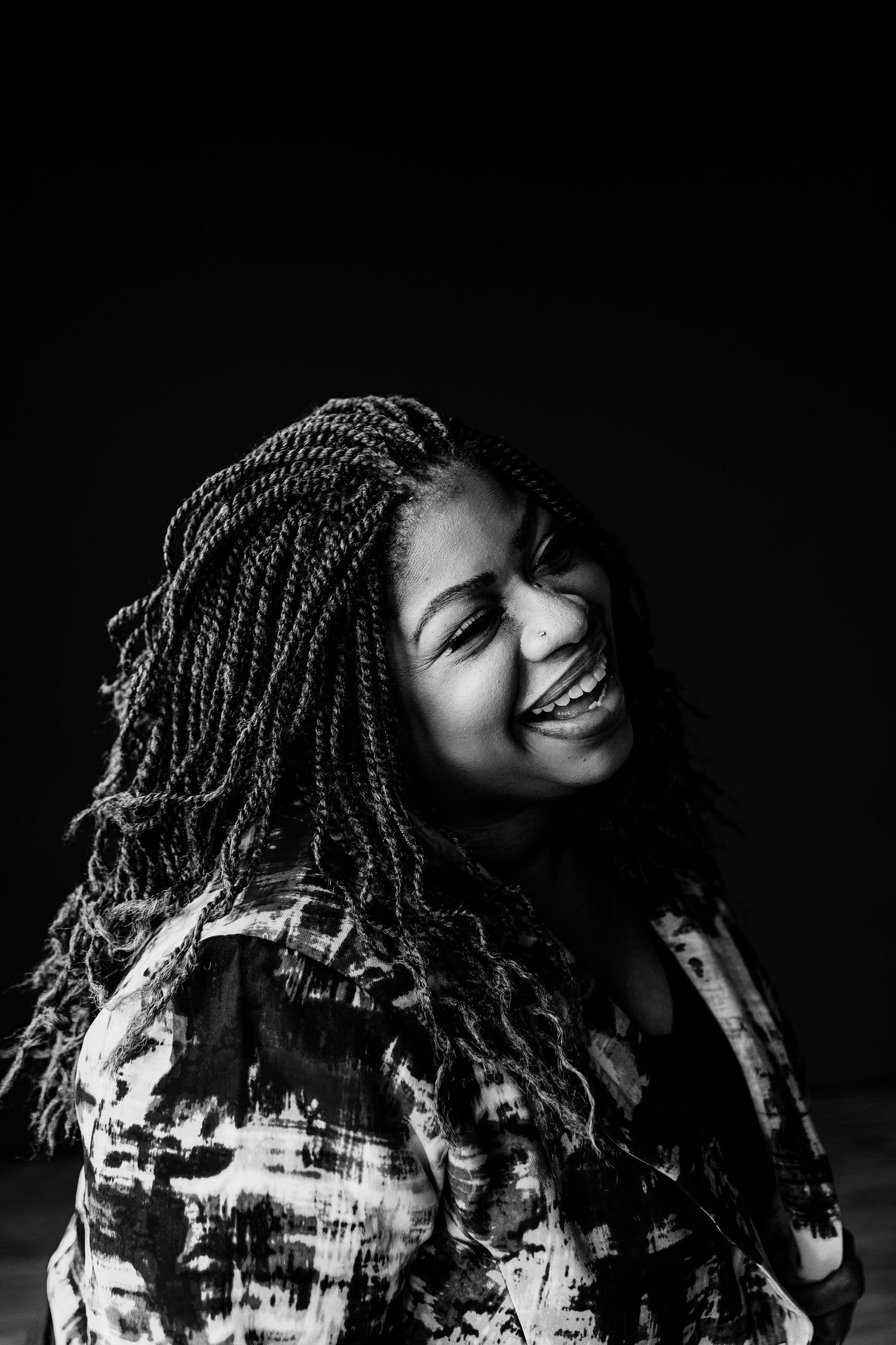 Black and white portrait of a laughing woman with long braided hair against dark background.