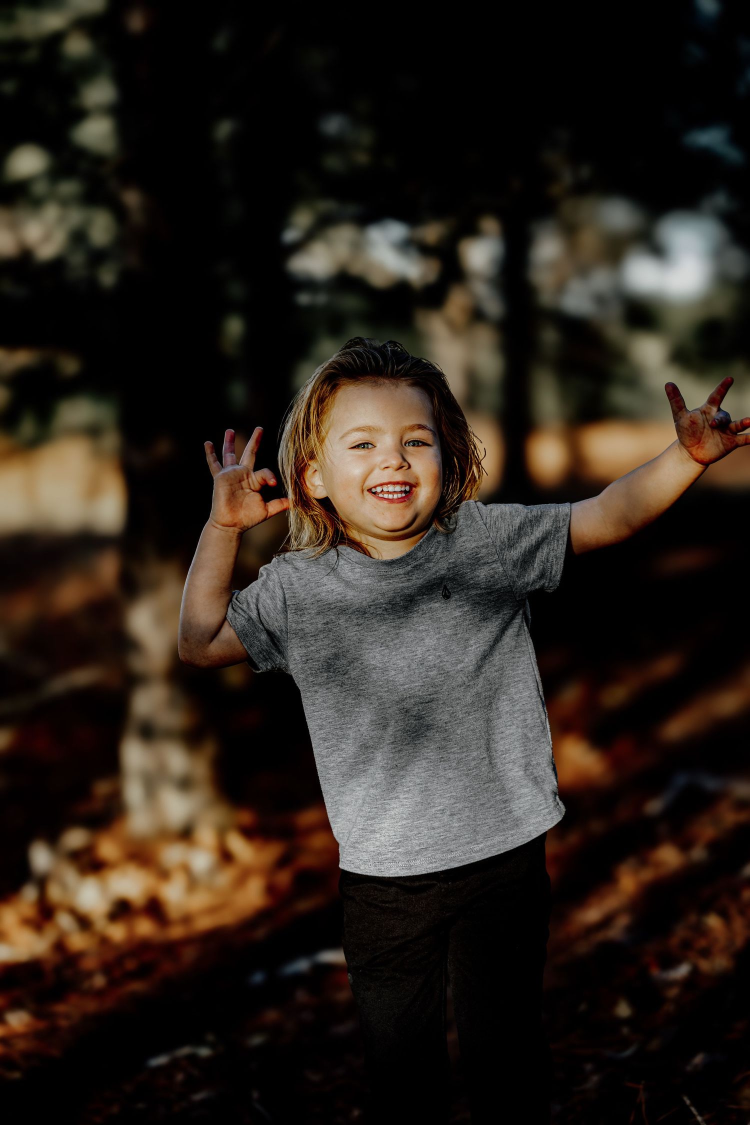Excited young child in gray tee making rock on hand gestures while laughing in autumn forest.