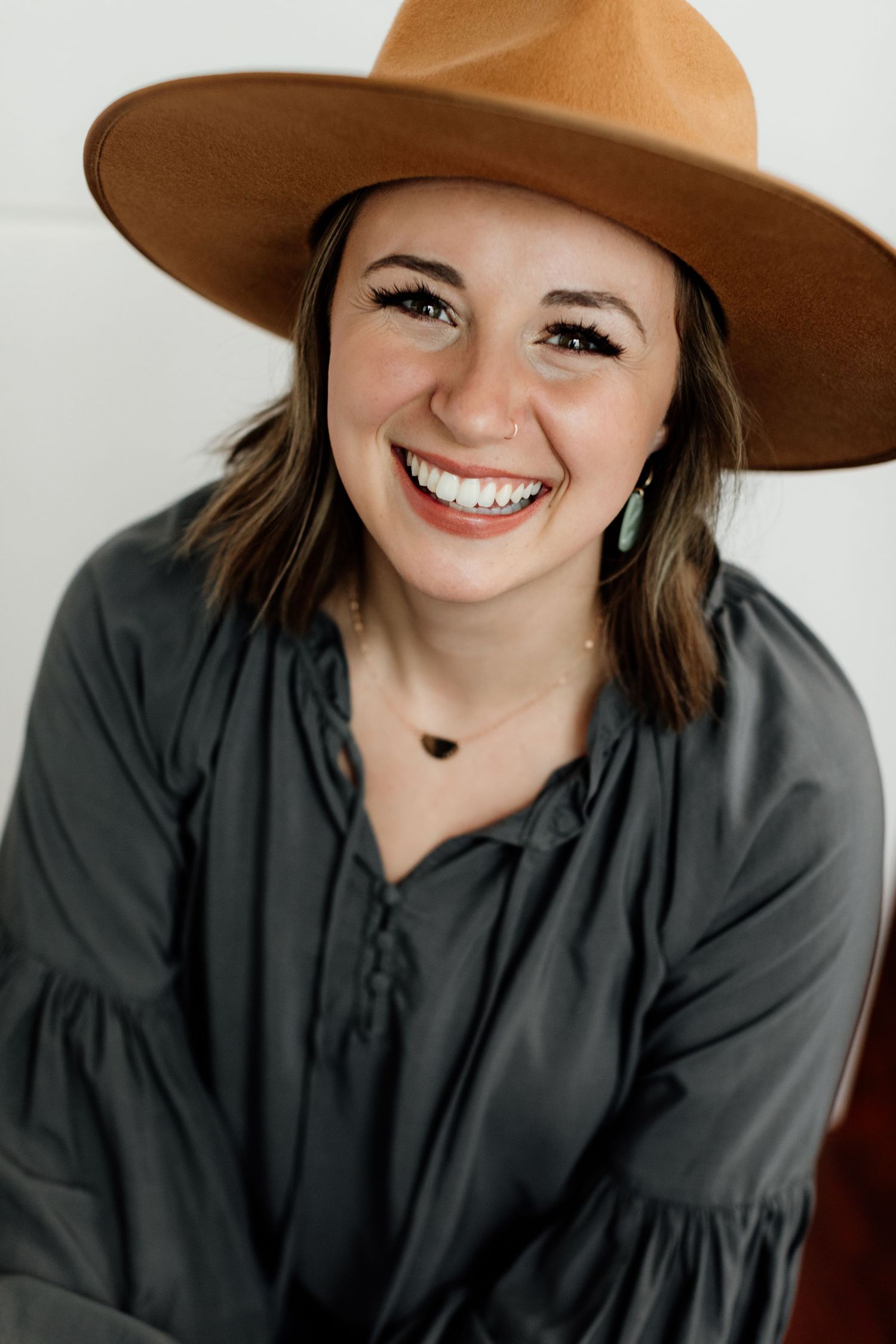 A cheerful young woman wearing a tan wide-brim hat and gray blouse smiling brightly.