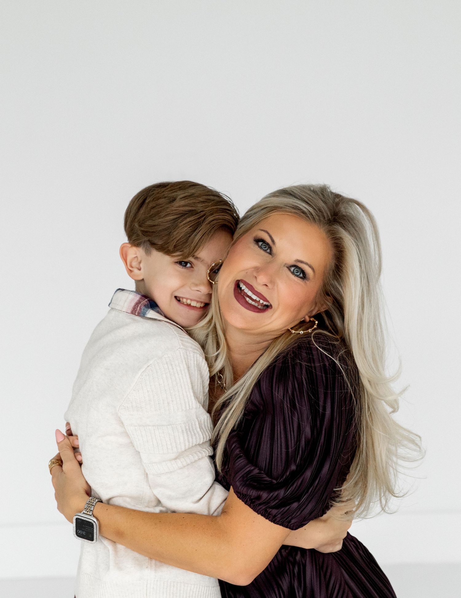 A woman with blonde hair hugs a smiling young boy in a white sweater against a light gray background.