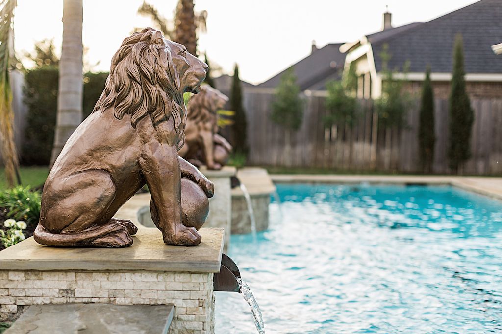 Bronze lion statue perched on stone ledge beside a sparkling backyard swimming pool with cypress trees in background.